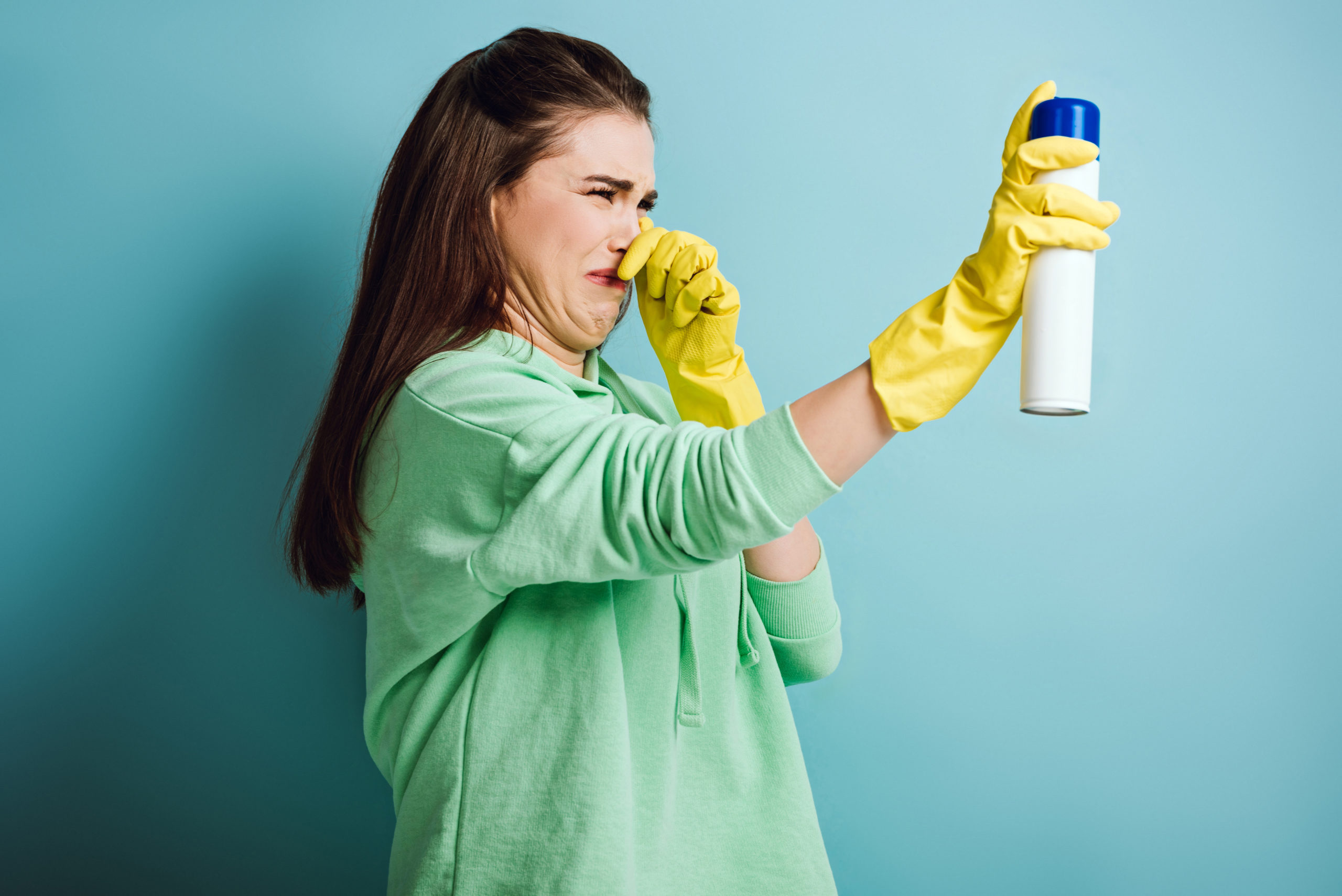 displeased housewife plugging nose with hand while spraying air freshener on blue background